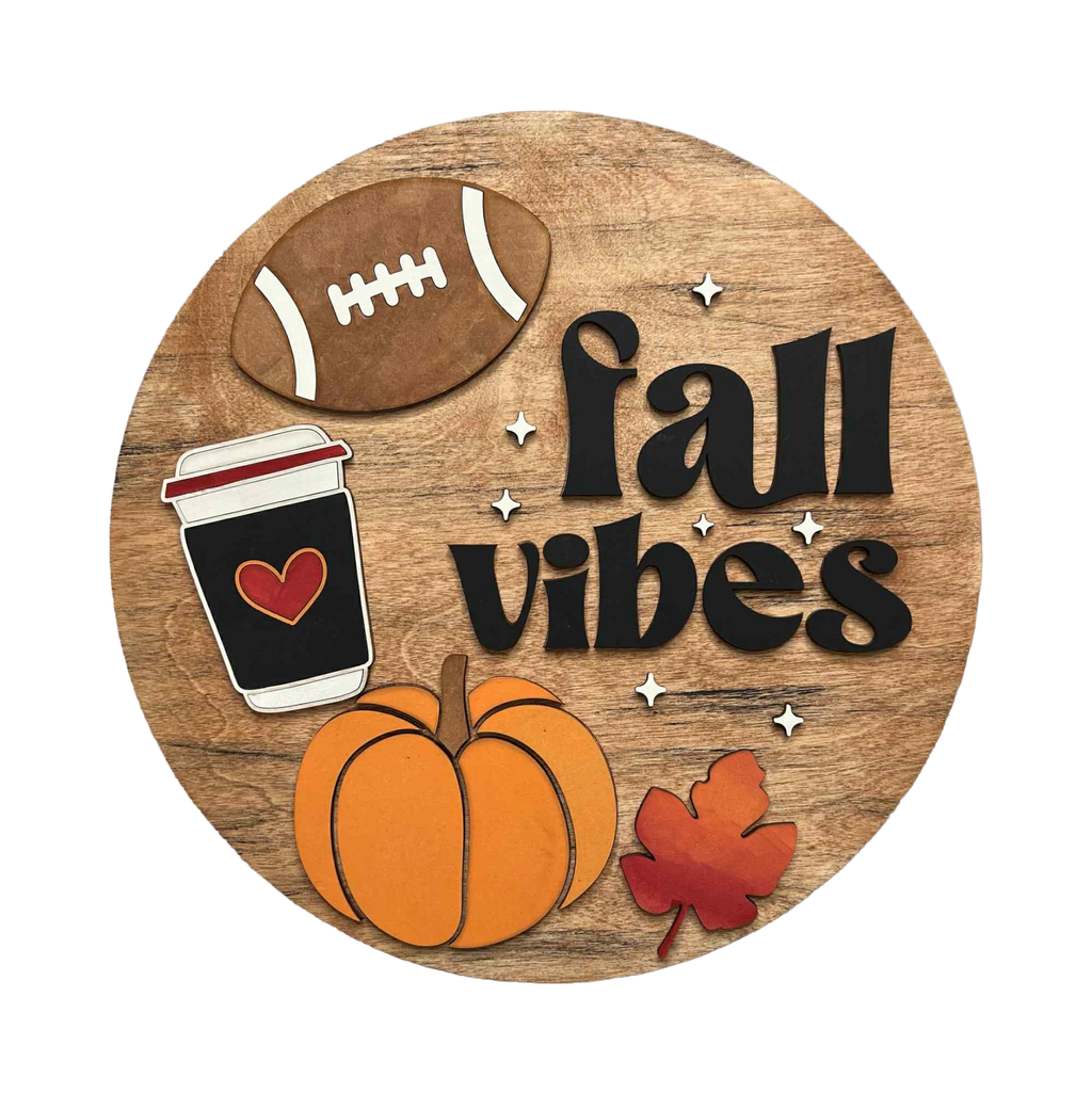 A circular wooden sign with a rustic wood-grain background featuring the words 'fall vibes' in black script, decorated with layered autumn-themed cutouts including a brown football, a black coffee cup with a red heart, an orange pumpkin, and a red maple leaf, available in San Antonio TX at DIY Sign Party.