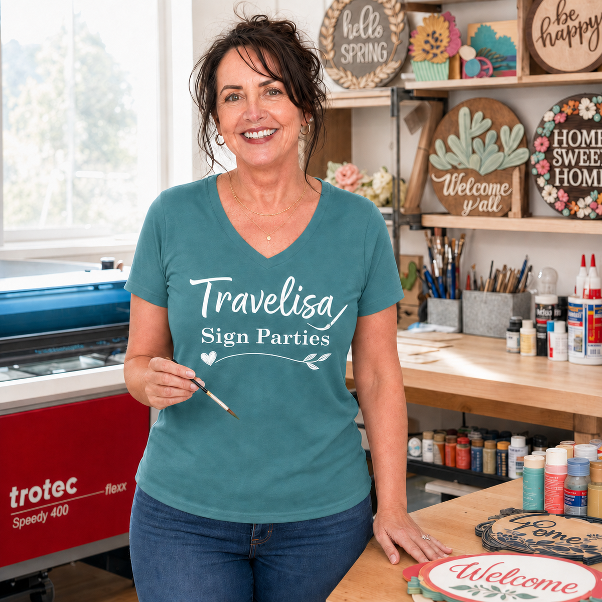 A smiling woman stands in a craft studio wearing a teal V-neck t-shirt with 'Travelisa Sign Parties' printed in white script. She is holding a paintbrush next to a wooden table with painted signs, with shelves of supplies and decorative signs in the background. Travelisa Sign Parties is located in San Antonio, TX. 