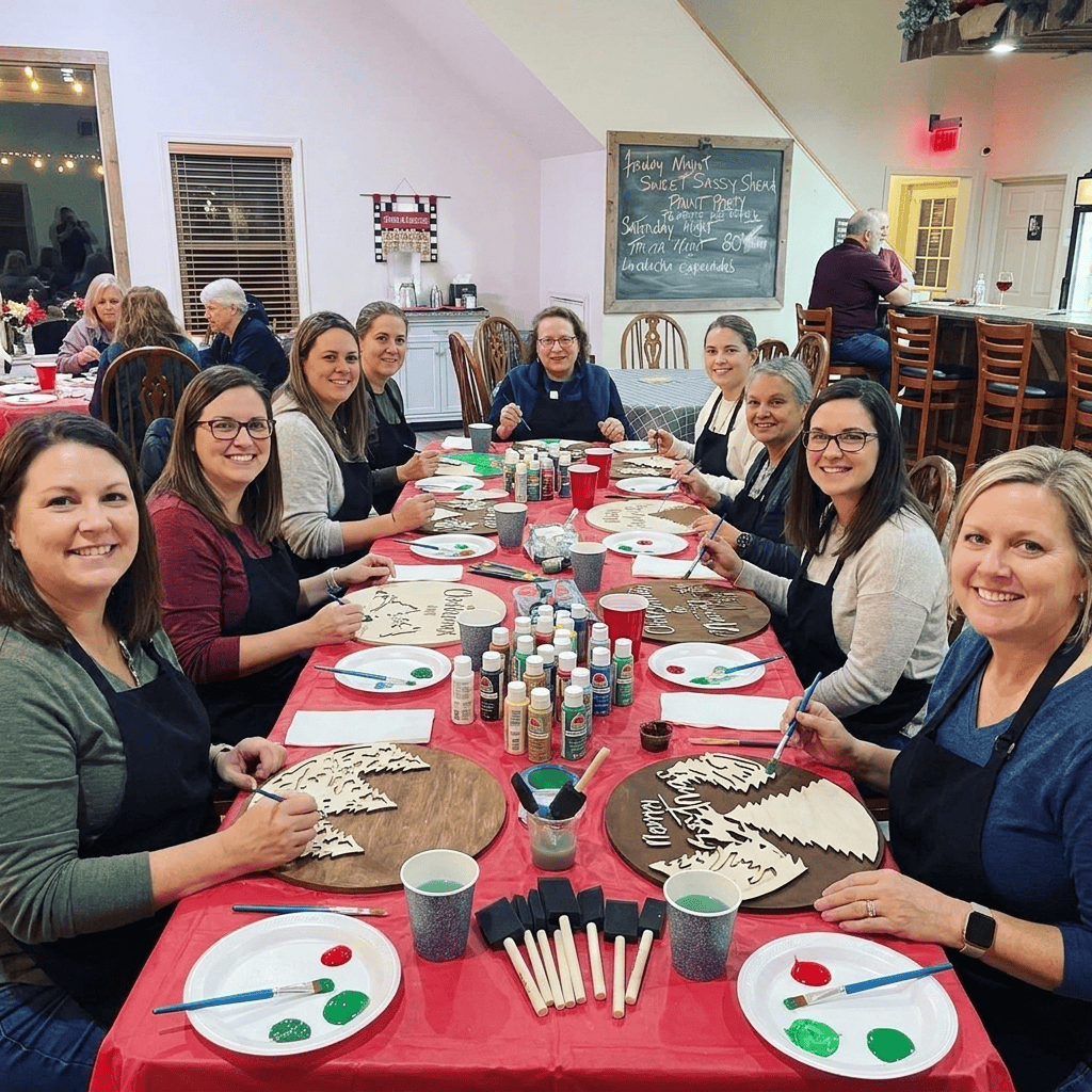A group of women sitting around a long table at a craft or painting workshop. The table is covered with paint supplies, including paint cans, brushes, sponges, and cups, with a red tablecloth. Ladies are smiling and having fun at a paint party in San Antonio, TX.